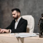 A businessman working on a laptop in an office with a Lady Justice statue in focus on the desk.